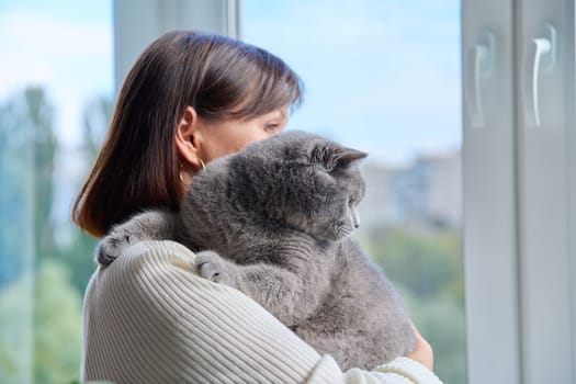 Woman hugging purring grey cat, holding pet in her arms, looking out the window together. Love, friendship, lifestyle, animals people concept