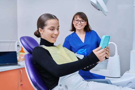 Happy young teenage female with mirror sitting in dentist chair smiling looking at healthy teeth, with dentist doctor in office. Dentistry, hygiene, treatment, dental health care concept