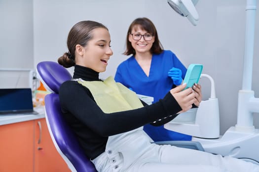 Happy young teenage female with mirror sitting in dentist chair smiling looking at healthy teeth, with dentist doctor in office. Dentistry, hygiene, treatment, dental health care concept