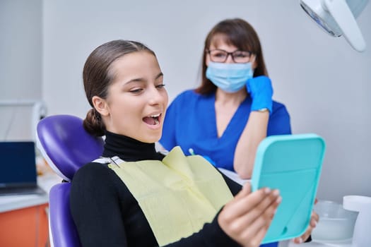 Happy young teenage female with mirror sitting in dentist chair smiling looking at healthy teeth, with dentist doctor in office. Dentistry, hygiene, treatment, dental health care concept