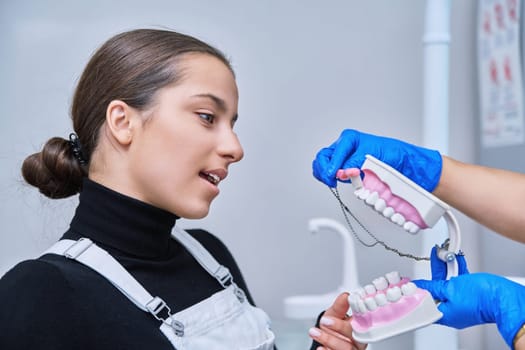 Teenage female sitting in dental chair at dentist checkup, doctor with dental jaw model and toothbrush telling and showing teenager dental care. Adolescence hygiene treatment dental teeth health care