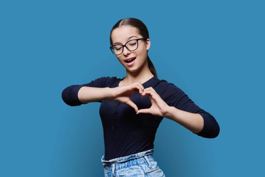 Young smiling female showing heart gesture with fingers, happy female showing love, blue studio background. Body language, signs symbols, love, emotions, romance youth concept