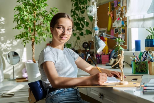 Portrait of teenage student sitting at desk home looking at camera. Smiling creative girl teenager 15, 16 years old with pencil drawing sketches. Education creativity leisure hobby adolescence concept