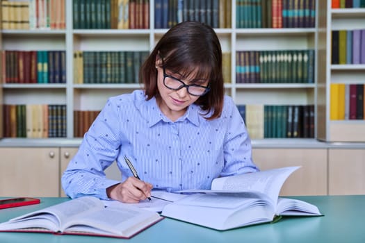 Middle-aged female teacher working in library, writing in papers at desk. Education, knowledge, school, college, university, technology, teaching concept