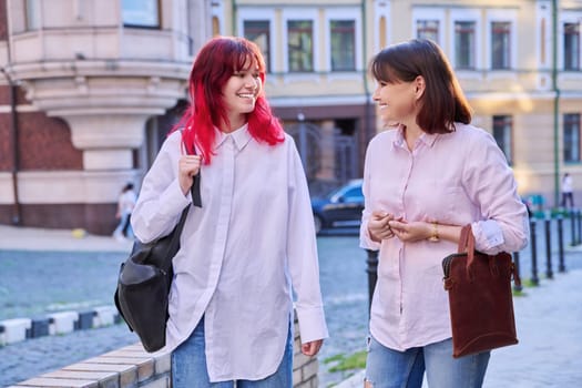 Two generations, smiling joyful happy mother and stylish attractive teenage daughter, walking talking together along city street. Family communication parent child teenager lifestyle leisure people