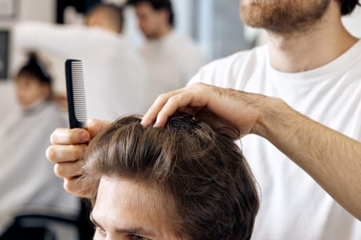 close-up, professional male hairstylist combing young customer's hair in barbershop