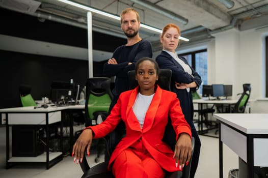 Caucasian red-haired woman, bearded Caucasian man stand behind a seated African American young woman in the office