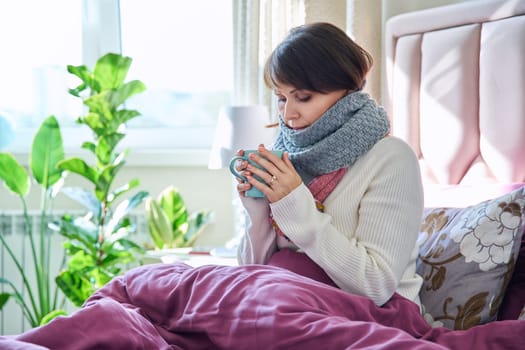 Middle aged woman sitting in bed under blanket warming herself with scarf and hot drink in mug, home lifestyle in cold autumn winter season