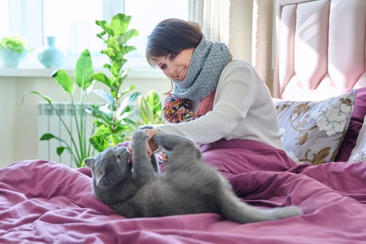 Middle-aged woman sitting at home in bed, warming with scarf and mug of hot drink, along with gray pet cat. Lifestyle, cozy, cold season autumn winter, comfort, animals, people concept