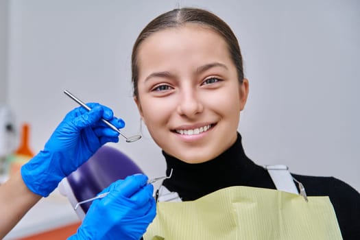 Portrait of young teenage girl in dental chair with hands of doctor with tools. Female teenager smiling with teeth looking at camera in dentist office. Adolescence hygiene treatment dental health care