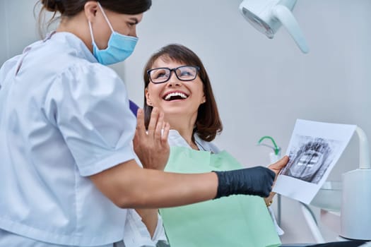 Female doctor dentist talking to middle aged woman patient in dental chair, discussing x-rays of teeth and jaws. Dental treatment and prosthetics, implantation, health care concept