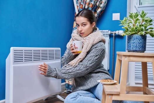 Cold autumn winter season, lifestyle, teenage female in warm sweater scarf warming near electric heating radiator, drinking hot drink with cup. Heating, warmth, equipment, life in cold season