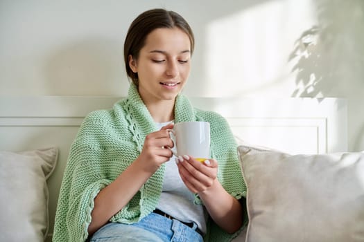 Young adolescent teenage girl resting basking under blanket with cup of hot tea, sitting on couch at home. Relax, rest, warming, cold season, lifestyle concept