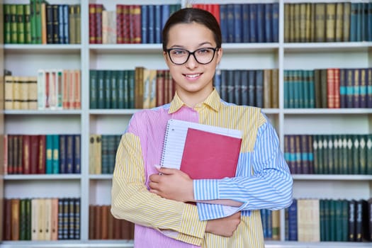 Portrait of teenager student girl looking at camera in library. Smiling teenage female 15, 16 years old wearing glasses, holding books. High school, education, knowledge, adolescence concept