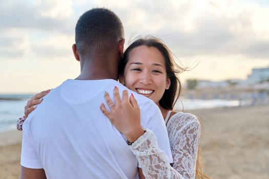 Young loving couple hugging on the beach. Face of beautiful Asian woman, man back. Love, relationship, honeymoon, vacation together, happiness, romantic, travel, family concept