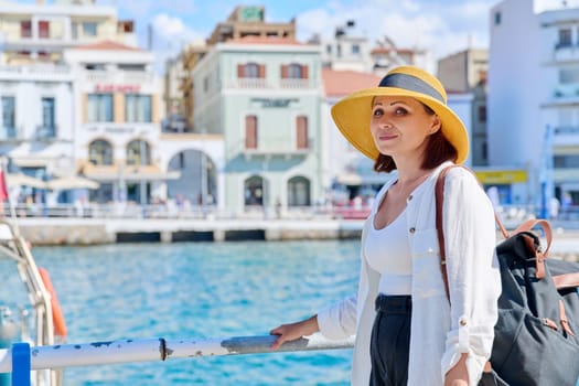 Beautiful mature woman in hat walking in seaside european city, copy space. Female tourist with backpack on the embankment on sunny summer day. Tourism, travel, vacation, people concept