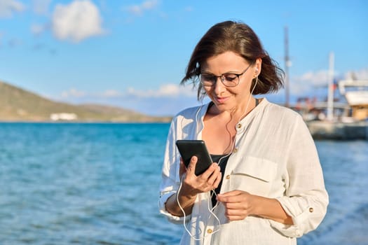 Happy 40s age woman in headphones with smartphone, sky sea nature. Beautiful female listening looking at smartphone screen walking relaxing on beach, copy space. Tourism, travel, vacation, technology