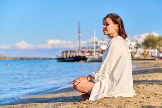Mature woman sitting in lotus position meditating on beach. Female enjoys sea nature, setting sun, summer vacations. Lifestyle, tourism, travel, beauty, health, relaxation, mature people concept