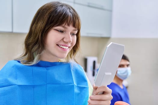 Teenage female looking at healthy teeth in mirror, in dental office, visit to dentist orthodontist. Treatment and care of teeth, teenagers, orthodontics, dentistry concept