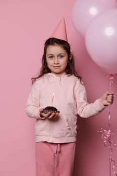 Beautiful lovely child, little girl dressed in pink clothes and festive party hat, holding bunch of inflatable pink pastel balloons and a birthday cake in her hands. Happy festive events. Celebrations