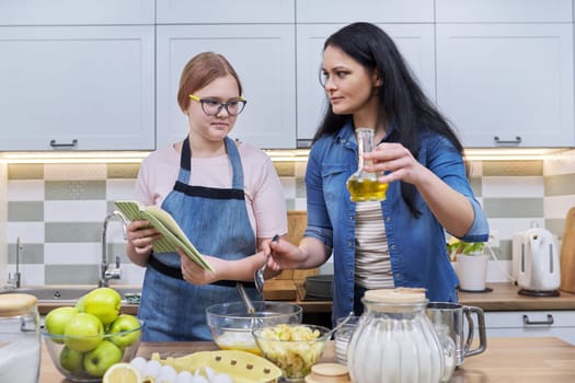 Mother and teenage daughter cooking at home in kitchen. Mom and girl making apple pie together, talking, use recipe book. Relationships, communication parent teenager, healthy homemade food, family