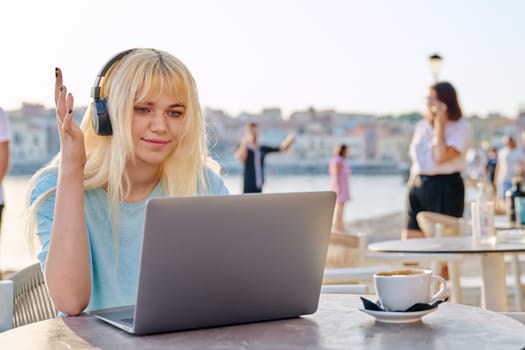 Smiling beautiful teenage girl in headphones looking into laptop. Blonde female in an outdoor cafe on waterfront using laptop. Youth, technology, lifestyle concept