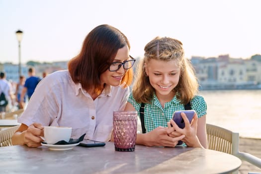 Mother and daughter child together in an outdoor cafe on the waterfront. Family, mom and preteen girl using smartphones, with coffee, drinks. Lifestyle, family, parent and child concept