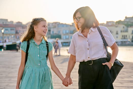 Mom and daughter child with a backpack holding hands together. Woman parent walking with child in city, outdoor. Generation, relationship, lifestyle, communication, family concept