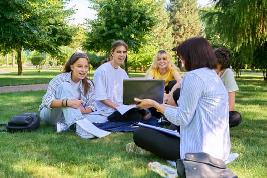 Outdoor, group of students with female teacher. Teenagers and mentor teacher talking sitting on grass in college park. Back to school, back to college, high school, education, teenagers concept