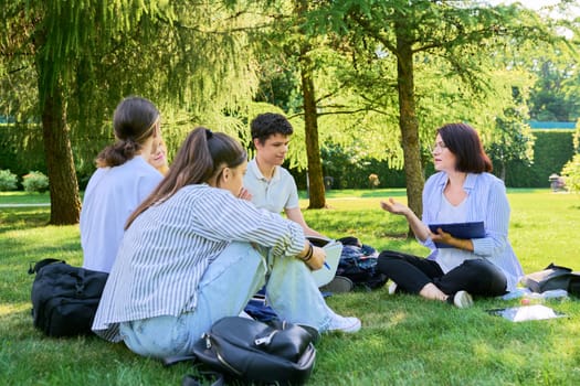 Outdoor, group of students with female teacher. Teenagers and mentor teacher talking sitting on grass in college park. Back to school, back to college, high school, education, teenagers concept