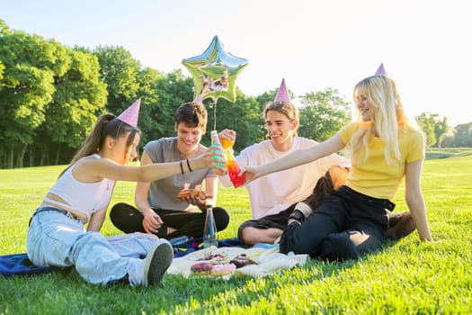 Birthday party. Teenager boy with cake with candles 17, celebrating birthday with friends, teenagers in festive hats sitting on grass in park, summer sunny day
