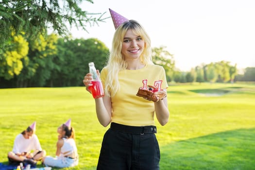 Beautiful teenage girl in festiv hat on birthday with cake and candles. Happy blonde female at outdoor picnic party holding cake with candles 17. Adolescence youth age beauty, holiday birthday concept