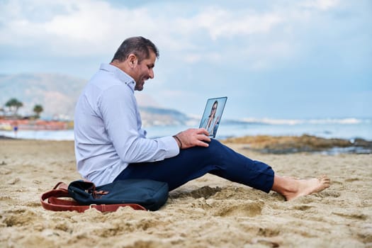 Online communication using wireless internet. Mature business man talking on webcam with female business partner using video call on laptop sitting outdoors on sandy sea beach