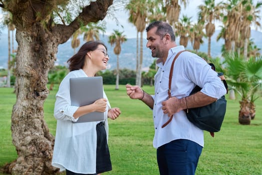 Middle aged business colleagues talking in the park. Mature man with backpack and woman with laptop meeting outdoors, positive smiling communicating