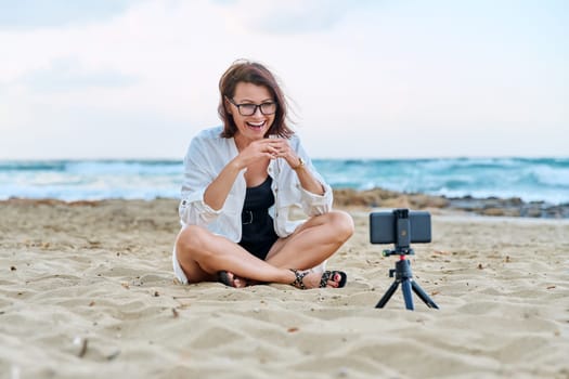 Middle aged woman sitting on beach with smartphone using video call. Female recording blog, vlog, talking online. Technology, telecommunications, lifestyle, freelance, training and business concept