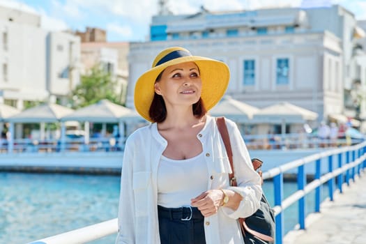 A beautiful mature woman in a hat walking along the seaside promenade of an old European tourist town. Summer, vacation, beauty, style, tourist travel, middle-aged people concept