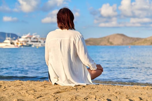 Mature woman sitting in lotus position meditating on the beach, back view. Female enjoys sea nature, setting sun, summer vacations. Lifestyle, tourism, travel, beauty, health, mature people concept