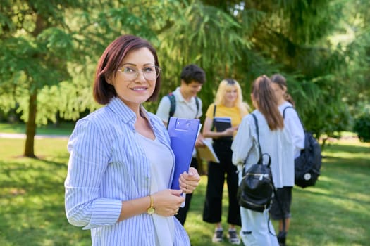 Middle aged female teacher mentor in glasses with digital tablet looking at camera, group of teenage students in park school college background. Education, teaching, profession, teachers day concept