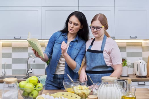 Mother and teenage daughter cooking at home in kitchen. Mom and girl making apple pie together, talking, use recipe book. Relationships, communication parent teenager, healthy homemade food, family