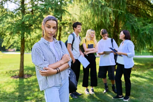 Female student 14, 15 years old with textbooks backpack, group of teenagers talking with teacher in park background. Education, teenage students, adolescence, school concept