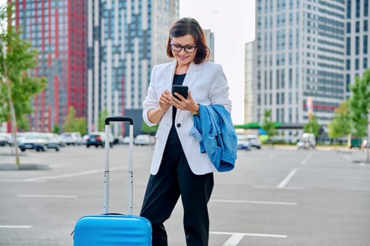 Middle aged confident business woman with suitcase, urban background. Smiling mature woman with luggage, looking at smartphone. Business trip, travel, city, work, lifestyle, people 40s concept