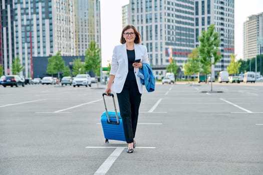 Mature confident business woman walking with suitcase, city background. Smiling middle agged woman with smartphone luggage, looking at camera. Business trip, travel, city, work, lifestyle, people 40s