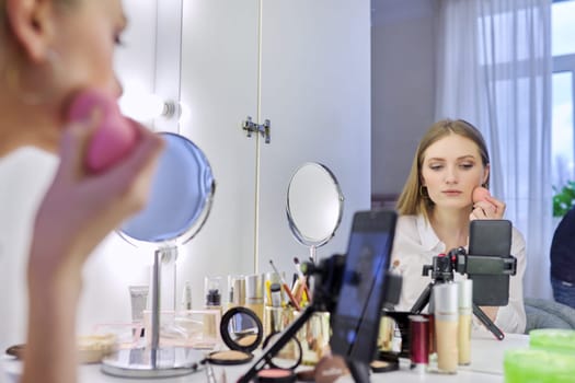 Young woman recording video, makeup beauty blog using smartphone. Female sitting in front of mirror, talking about cosmetics and showing application. Beauty, fashion, care, technology, youth concept