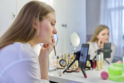 Young woman recording video, makeup beauty blog using smartphone. Female sitting in front of mirror, talking about cosmetics and showing application. Beauty, fashion, care, technology, youth concept