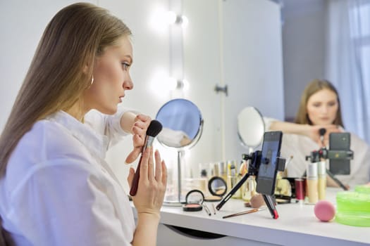 Young woman recording video, makeup beauty blog using smartphone. Female sitting in front of mirror, talking about cosmetics and showing application. Beauty, fashion, care, technology, youth concept