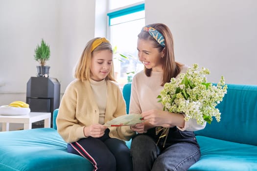 Happy mother and daughter child congratulating with bouquet of flowers and hand-drawn card, at home in living room on sofa. Mother's day, family, relationship, emotions, love, togetherness concept
