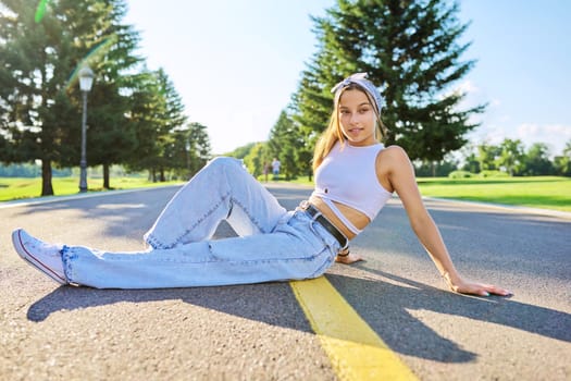 Fashionable teenage girl posing sitting on the road in park. Female hipster teenager wearing trendy outdoor headband. Fashion, style, beauty, youth concept