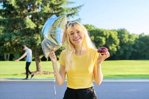 Portrait of cheerful beautiful teenage blonde with cake donut and silver balloon, in sunny park. Beauty, fashion, style, youth, vacation, summer, birthday, holiday, joy, happiness, adolescence concept