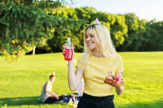 Beautiful teenage girl in festiv hat on birthday with cake and candles. Happy blonde female at outdoor picnic party holding cake with candles 17. Adolescence youth age beauty, holiday birthday concept