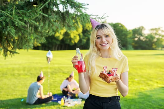 Beautiful teenage girl in festiv hat on birthday with cake and candles. Happy blonde female at outdoor picnic party holding cake with candles 17. Adolescence youth age beauty, holiday birthday concept